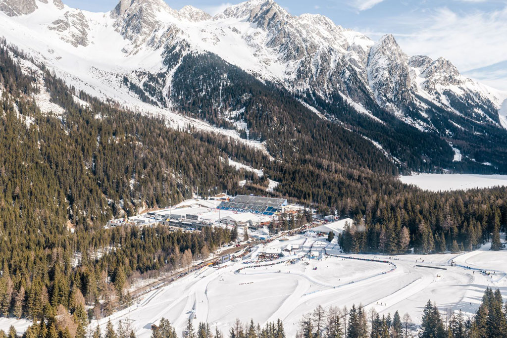 Luftaufnahme eines Wintersportstadions mit Loipen und Tribünen, eingebettet in einen verschneiten Nadelwald. Im Hintergrund ragen steile, schneebedeckte Berggipfel unter einem blauen Himmel auf.