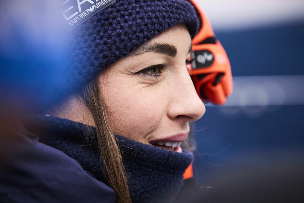 Nahaufnahme der italienischen Biathletin Dorothea Wierer mit dunkelblauer Wollmütze und Winterjacke. Sie lächelt leicht und blickt konzentriert zur Seite. Im Hintergrund ist unscharf ein orangefarbenes Fernglas oder ein Sportgerät zu erkennen.